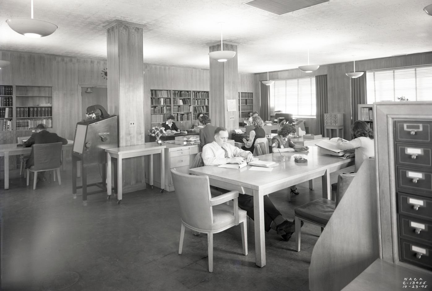 Several people using a library. 
Many are seated at tables reading books. 
There are book shelves, file cabinets for index cards, 
and a microfische reader.
